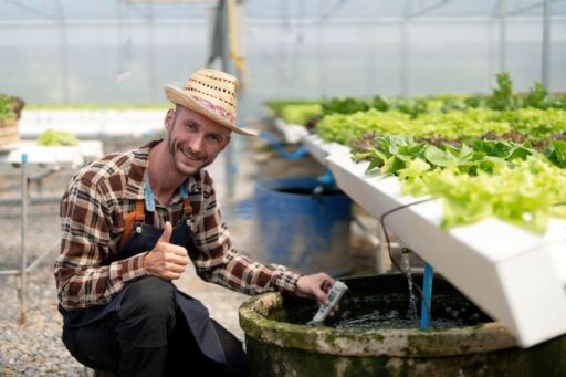 Agricultor sonriendo mientras mide el pH y la calidad del agua en un tanque de peces con un medidor digital dentro de un invernadero acuap&oacute;nico.